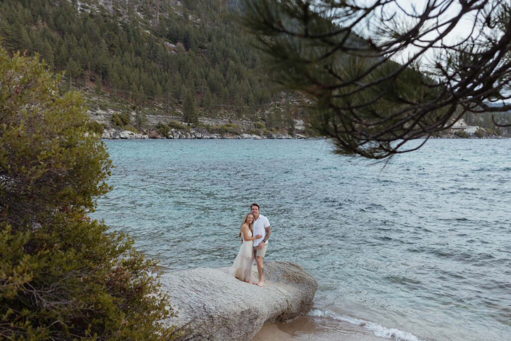 Engagement couple standing on large rock together and smiling at camera with Lake Tahoe in background