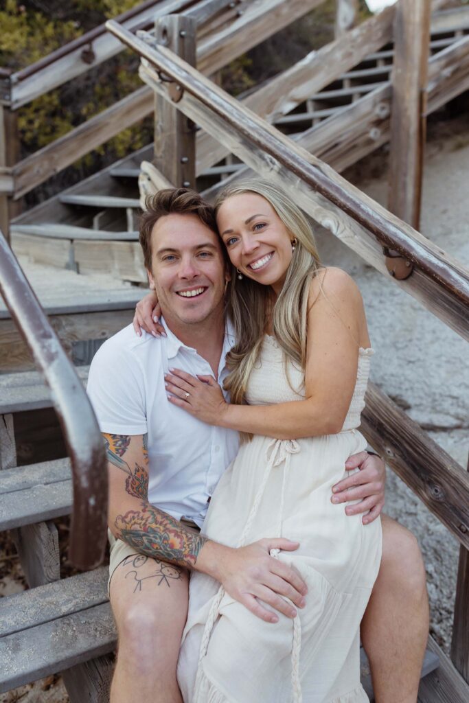 Engagement couple sitting on wooden staircase together and smiling at camera while on a beach in Lake Tahoe