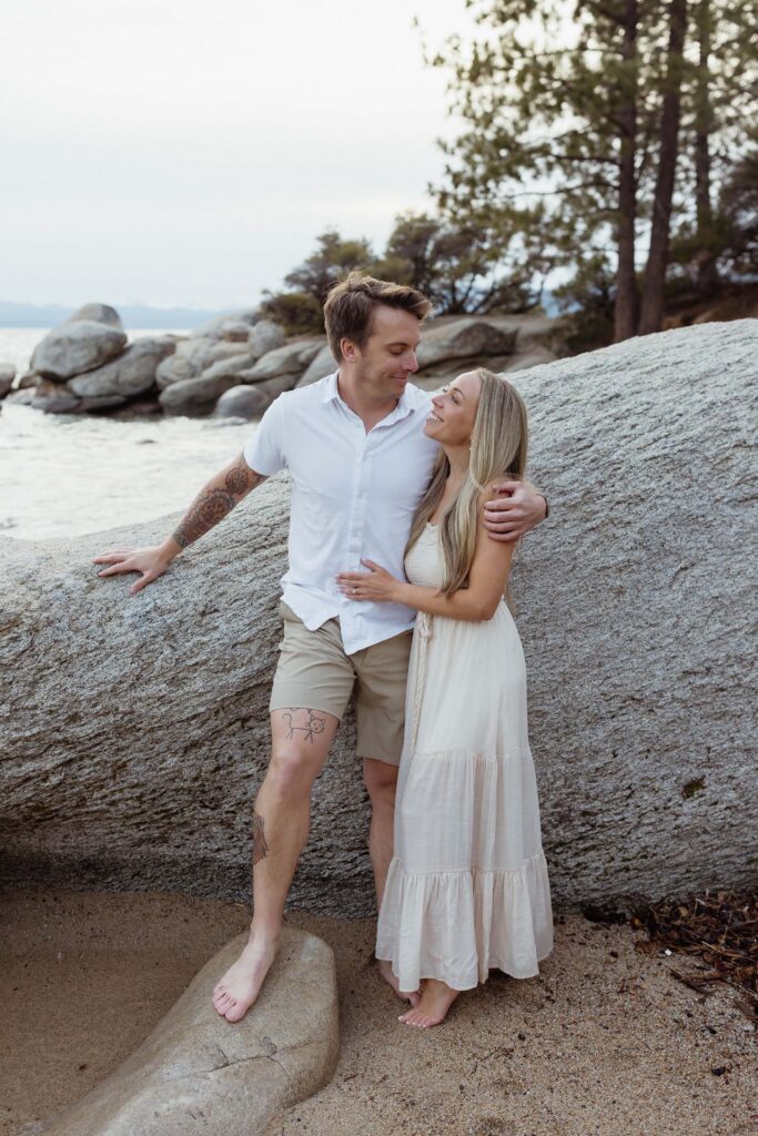Engagement couple smiling at each other while leaning on large rock together on a Lake Tahoe beach