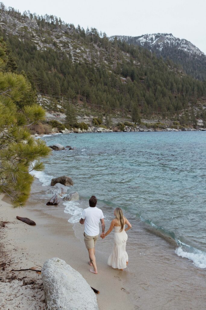 Engagement couple holding hands while walking along sandy beach next to Lake Tahoe  with lots of pine trees in background