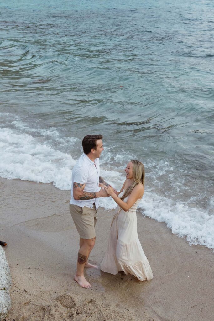 Engagement couple holding hands and laughing with each other while standing on a sandy beach next to Lake Tahoe together