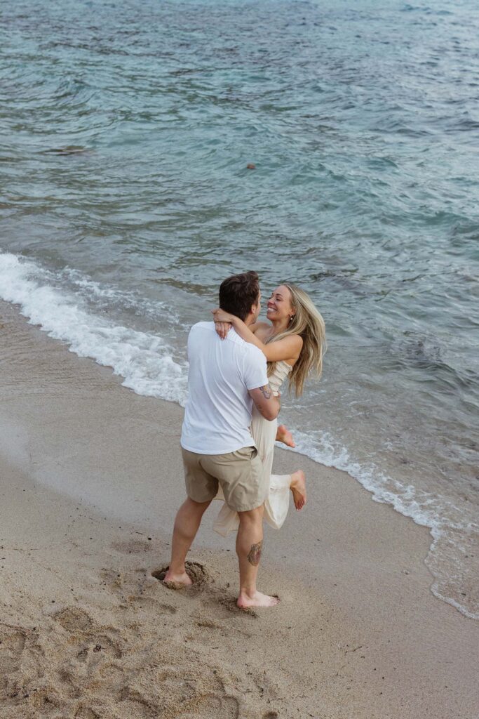 Man twirling fiancé around while she smiles on a sandy beach in Lake Tahoe