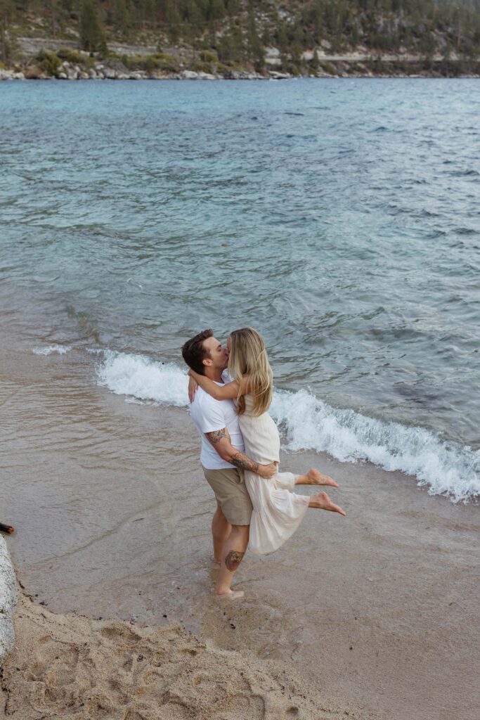 Man holding fiancé up and kissing her while standing barefoot on a sandy beach in Lake Tahoe with water in background