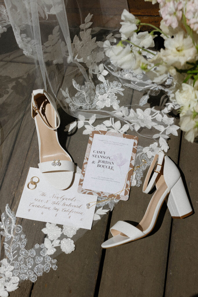 Wedding details in flat lay with announcement, invitations, bride's shoes, floral bouquet and wedding rings partially covered by veil on wooden deck at Garwoods Lake Tahoe