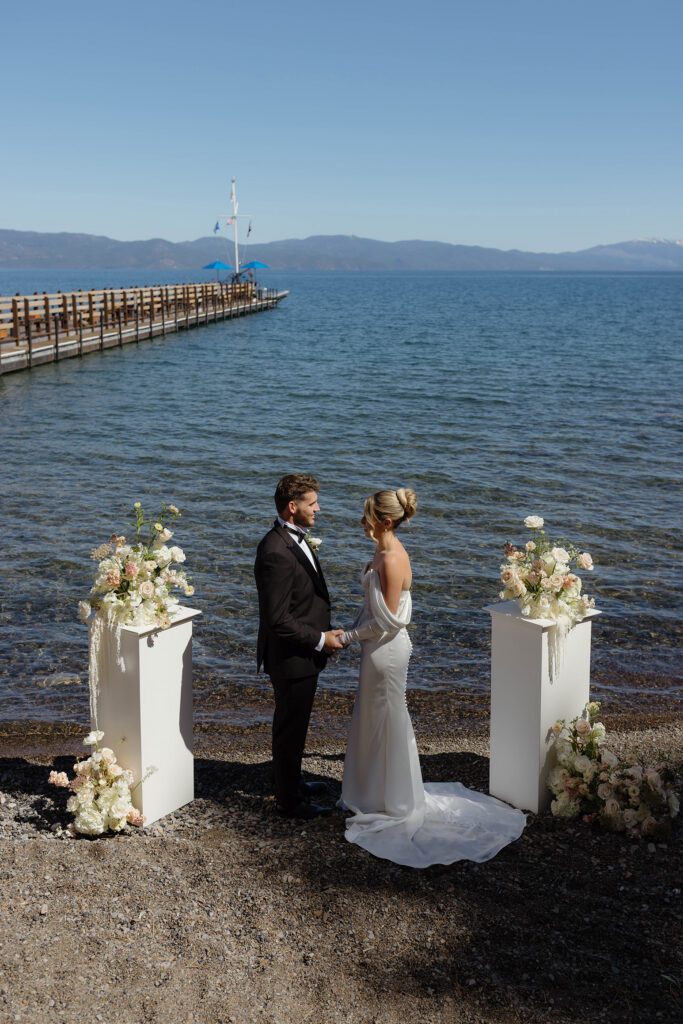 Wedding couple holding hands in between two white pillars with colorful florals during ceremony next to Lake Tahoe at Garwoods