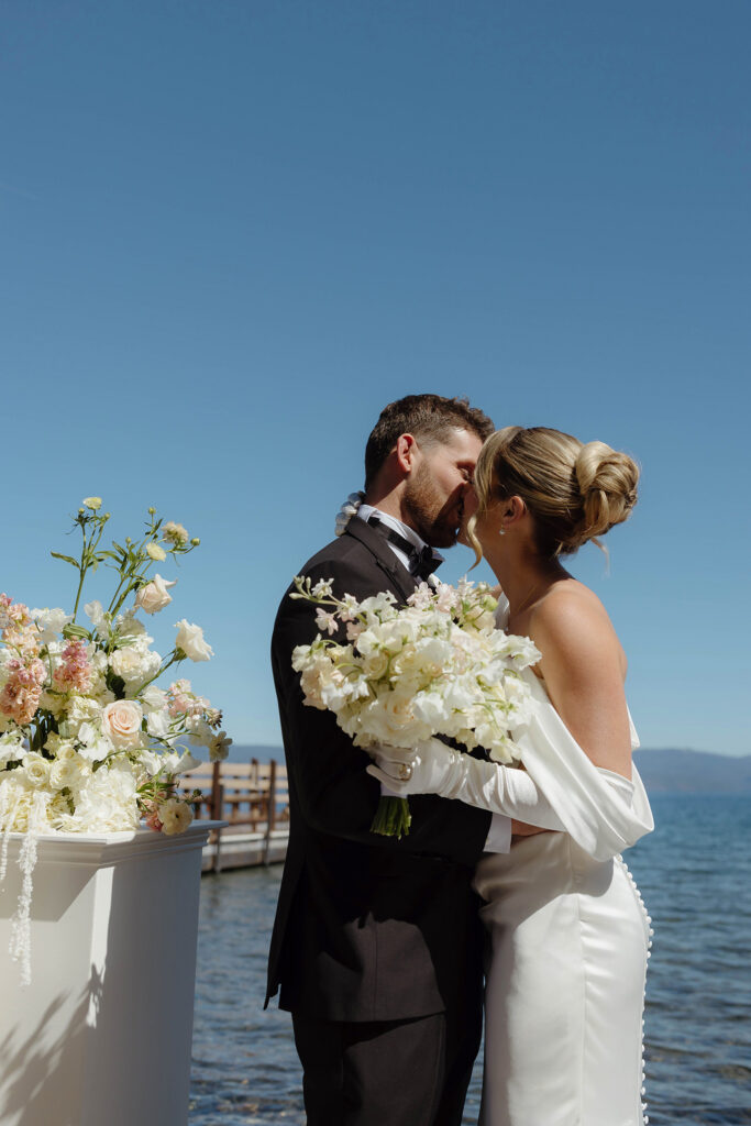 Wedding couple kissing during ceremony next to white pillar with florals and bride holds white floral bouquet next to Lake Tahoe
