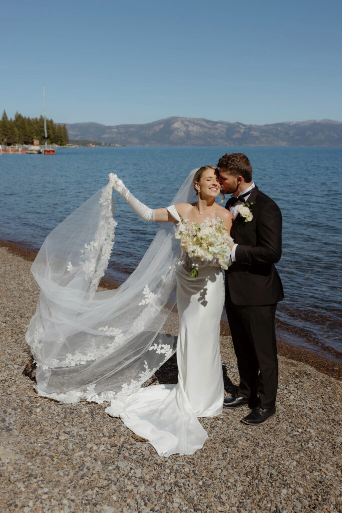 Wedding couple standing on the shore of Lake Tahoe while groom kisses bride's cheek and she holds her veil out to her side