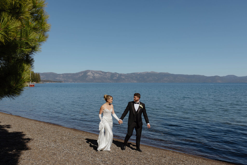 Wedding couple holding hands while smiling at each other and walking along shore next to Lake Tahoe