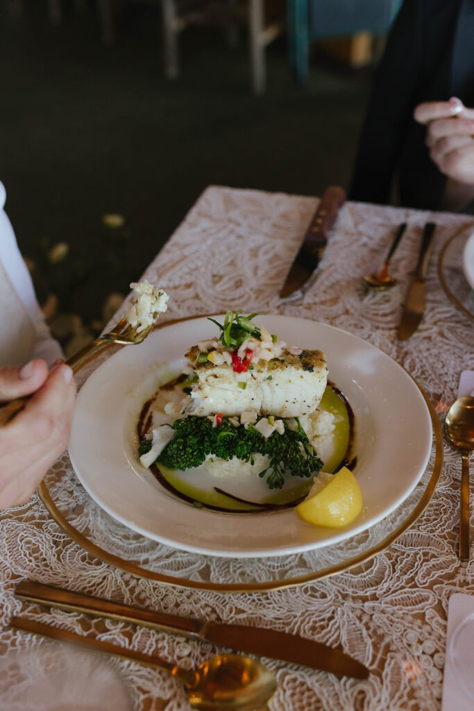 Bride eating halibut over broccolini while at wedding dinner table with lace white tablecloth and golden cutlery at Garwoods Lake Tahoe