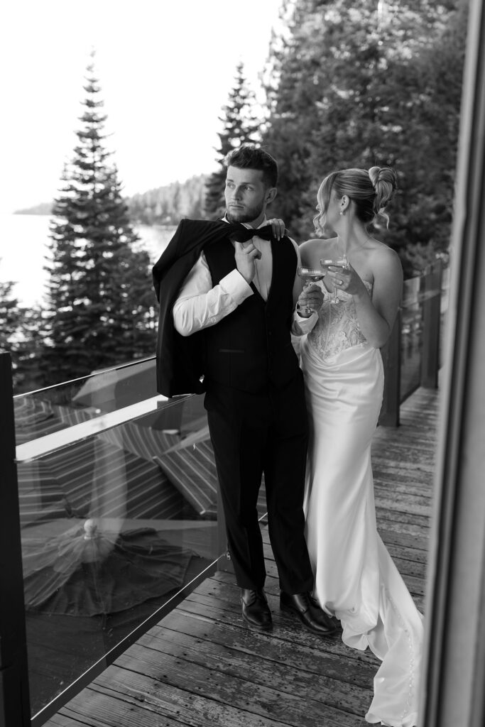 Wedding couple standing together on deck overlooking Lake Tahoe while holding drinks and groom has jacket draped over shoulder at Garwoods