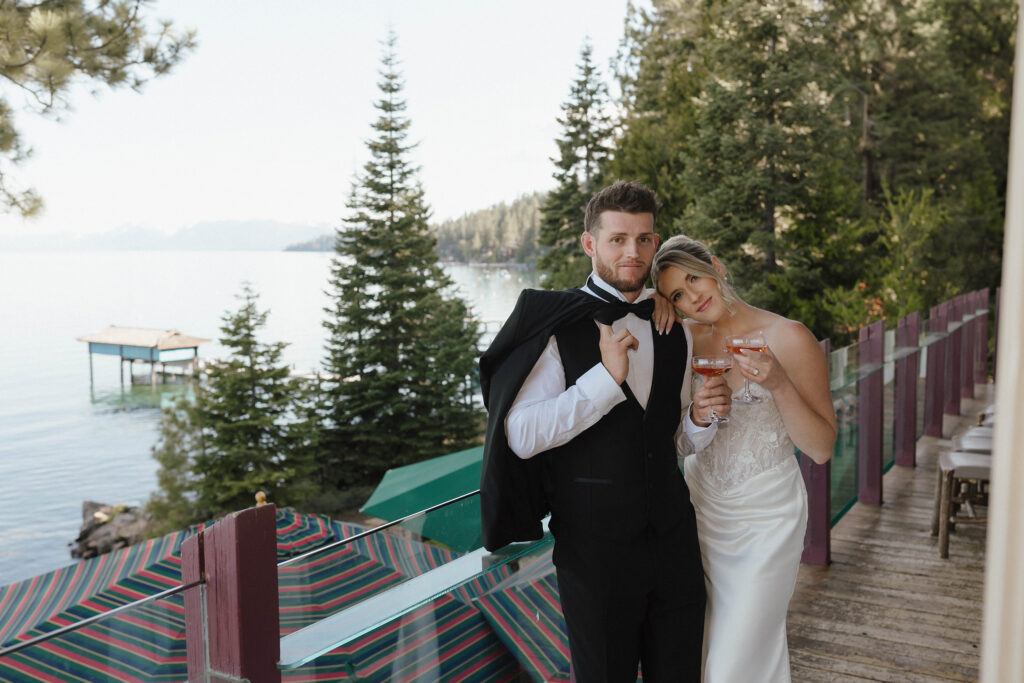 Wedding couple leaning on each other while smiling at camera and holding drinks on deck overlooking Lake Tahoe at Garwoods