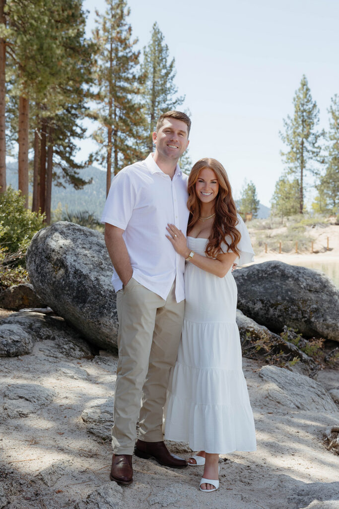 Engagement couple standing with arms around each other and smiling at camera on sand harbor beach with large rocks behind them