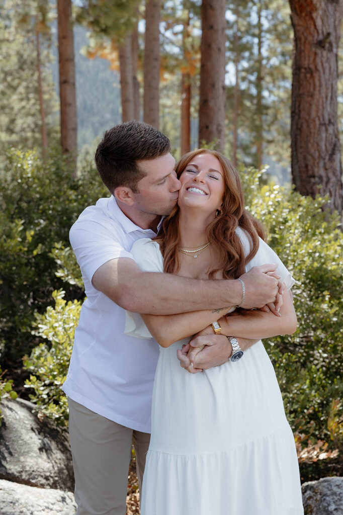 Man kissing fiancé on cheek and wrapping arms around her while she smiles at Sand Harbor with bushes and trees in background