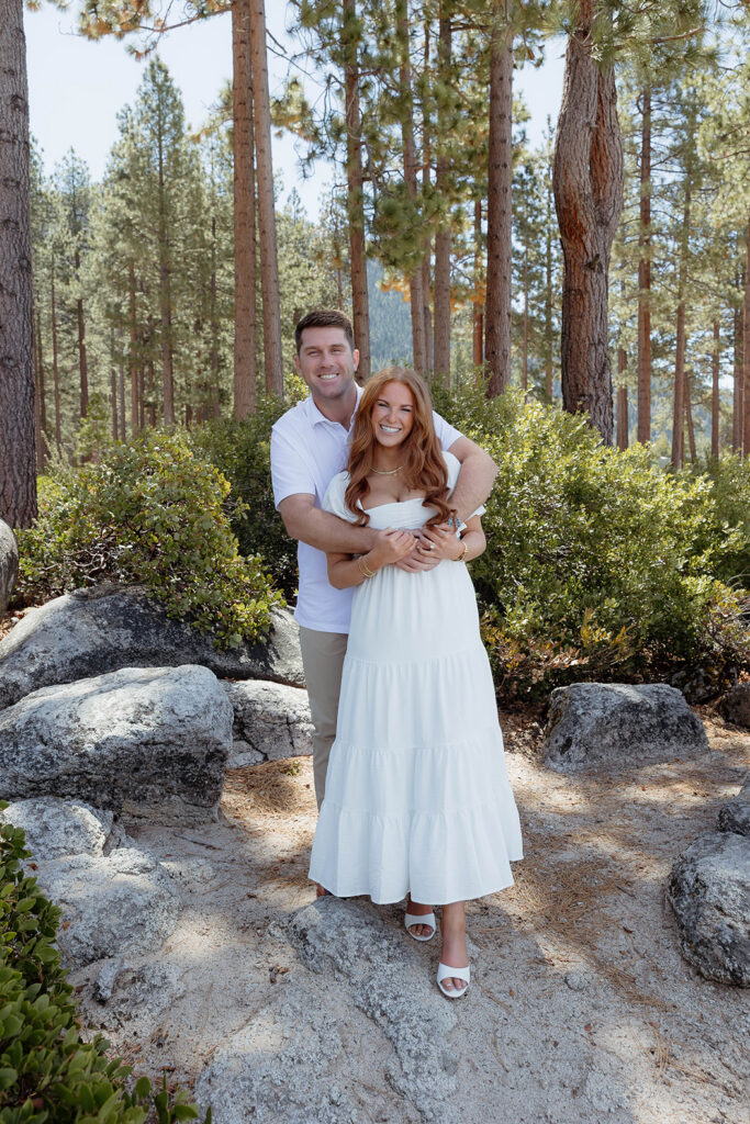 Man hugging fiancé from behind while both smile at the camera in Sand Harbor with tall trees and bushes in backgorund