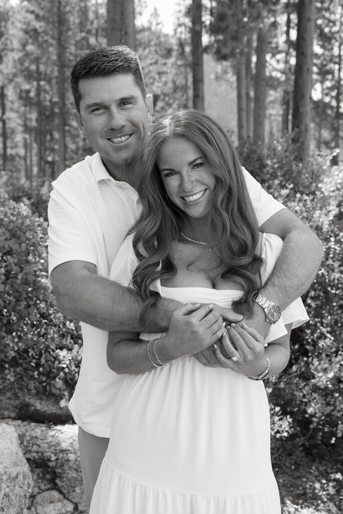 Close up of man with arms wrapped around fiancé while both smile at camera in Sand Harbor with trees in background