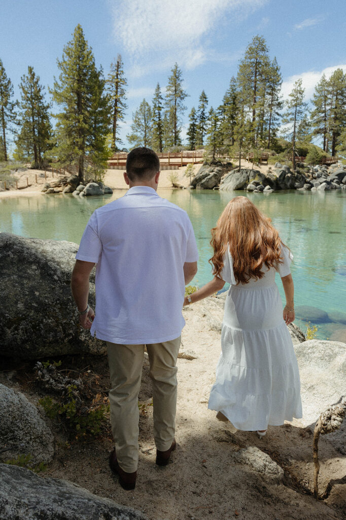 Engagement couple holding hands while walking down to sunny beach at Sand Harbor with tall trees in background