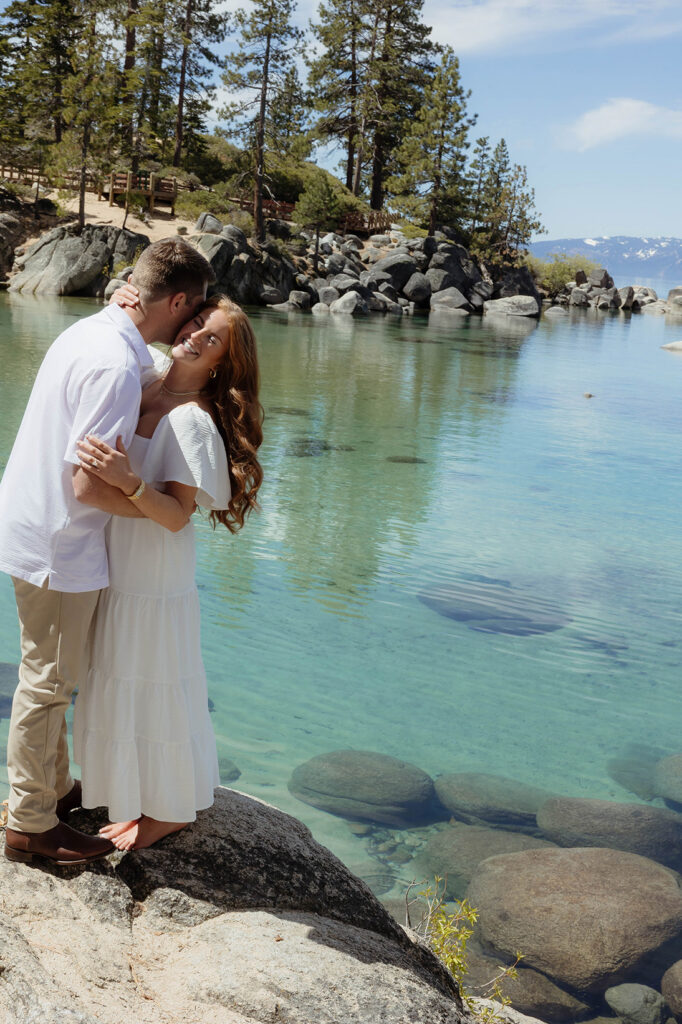 Engagement couple holding each other while standing on a rock next to Lake Tahoe at Sand Harbor with pine trees in background