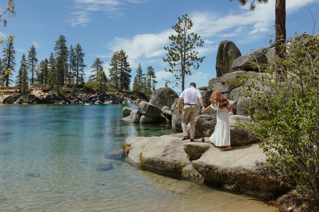 Engagement couple holding hands while walking across large rocks together next to Lake Tahoe at Sand Harbor