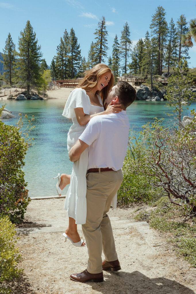 Man lifting fiancé up while she laughs and holds his neck with Lake Tahoe in background at Sand Harbor