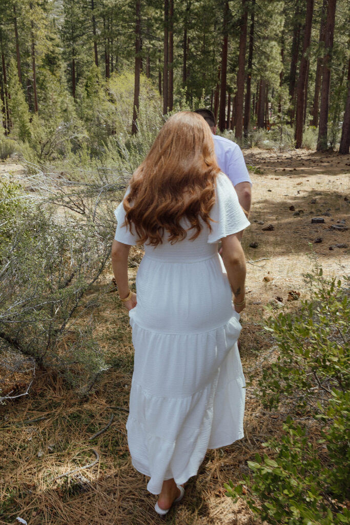 Woman holding dress as engagement couple walks through trail together at Sand Harbor towards tall pine trees