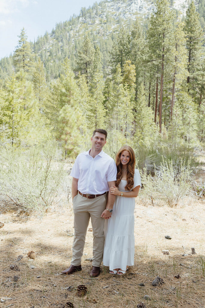 Engagement couple standing next to each other and holding hands while smiling at camera with ground covered in pine needles and tall trees in background at Sand Harbor