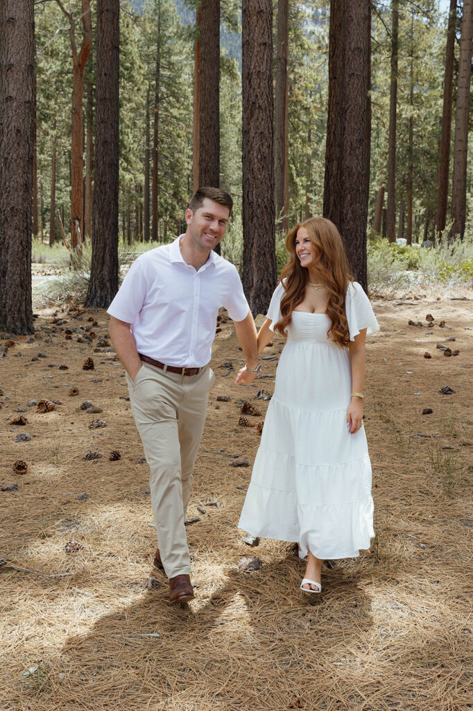 Engagement couple holding hands while smiling and walking across pine needle covered ground with tall pine trees behind them at Sand Harbor