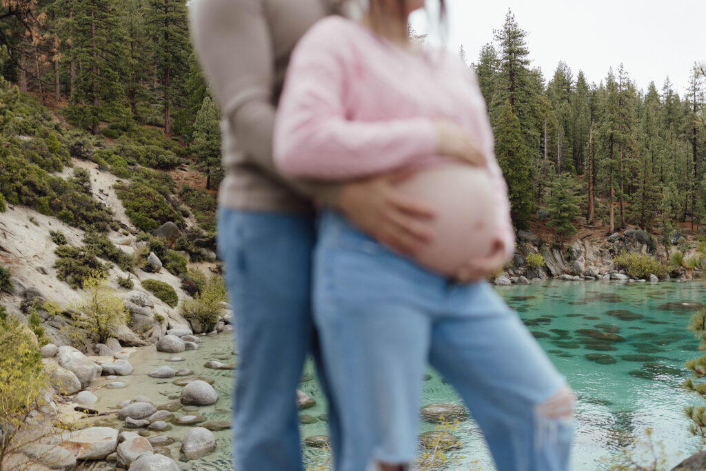 Close up of couple holding pregnant woman's belly with sandy beach and large rocks in background at Lake Tahoe