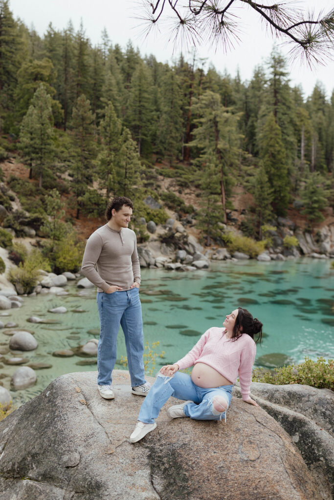 Man standing and looking down at pregnant wife sitting while both are on large rock with Lake Tahoe in background