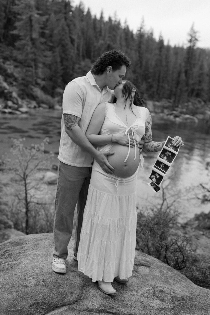 Couple kissing and standing on large rock together while holding pregnant woman's belly and maternity photos with Lake Tahoe in background