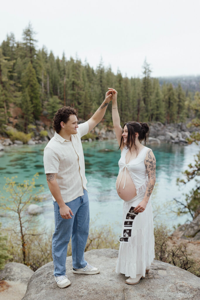 Couple holding hands together in the air while woman shows pregnant belly and holds maternity photos and smiling with Lake Tahoe in background