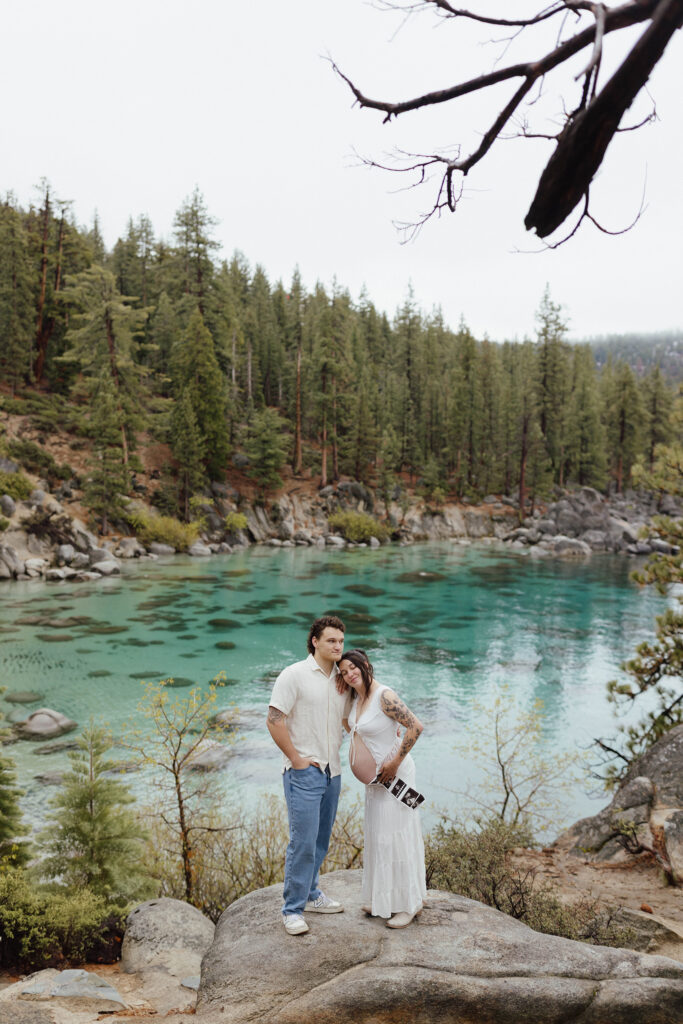 Couple standing next to each other on large rock while woman shows pregnant belly and holds maternity photos with lots of pine trees and Lake Tahoe in background