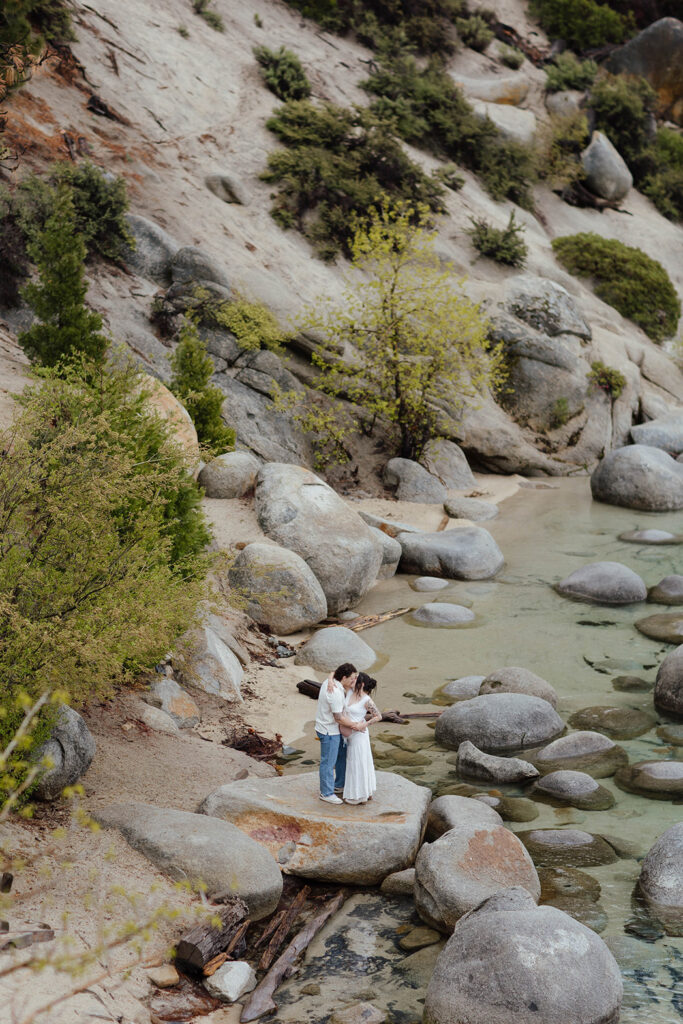 Couple standing on large rock and holding each other while kissing on a sandy beach in Lake Tahoe