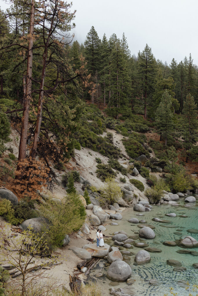 Couple in the distance holding each other while standing on a large rock on a beach in Lake Tahoe