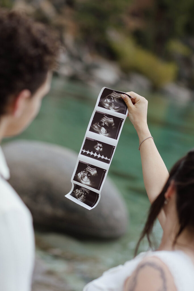 Woman holding up maternity photos while couple looks at them with Lake Tahoe and large rocks in background