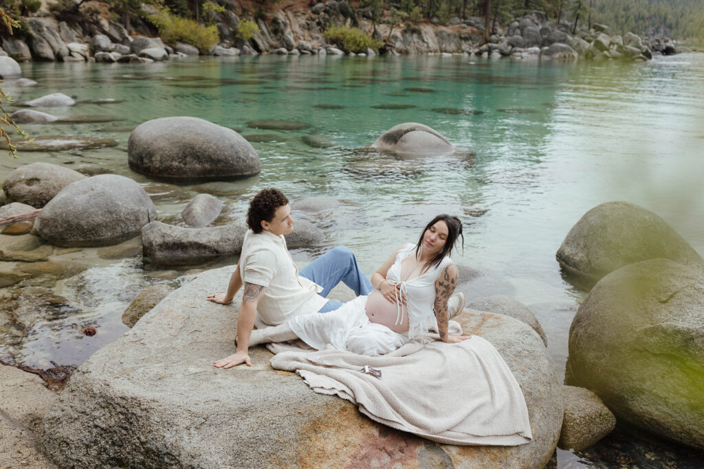 Couple leaning back on a boulder together while woman holds pregnant belly next to Lake Tahoe