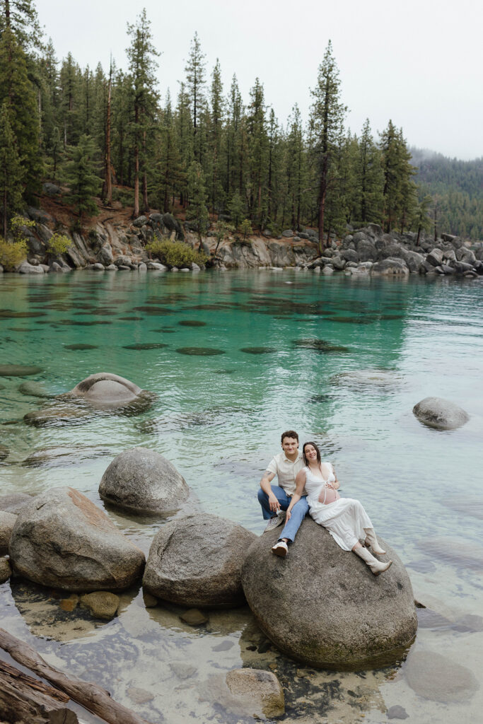 Couple sitting on large rock on Lake Tahoe beach together while smiling at camera and woman holds her pregnant belly