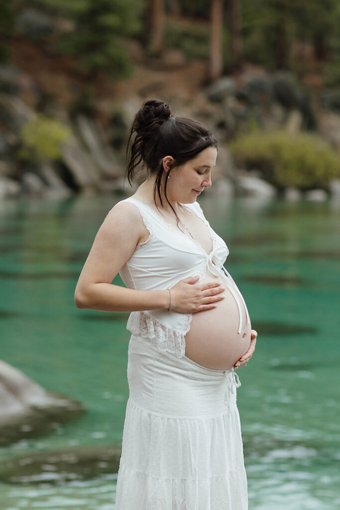 Woman in white dress holding her exposed pregnant belly and looking down with Lake Tahoe in background