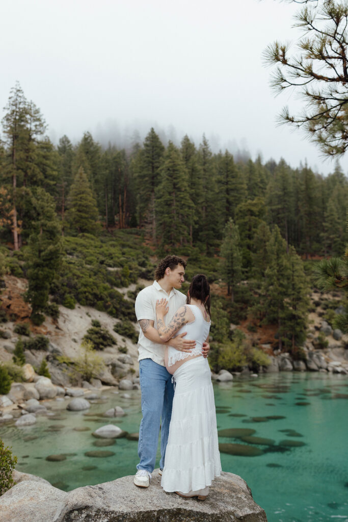 Couple holding each other while standing on large rock together overlooking Lake Tahoe beach with lots of pine trees in background