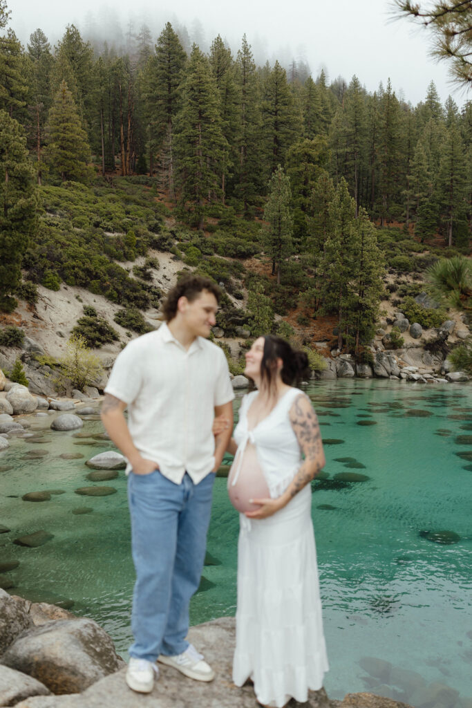 Couple smiling at each other while standing on large rock over looking Lake Tahoe beach and woman holds pregnant belly