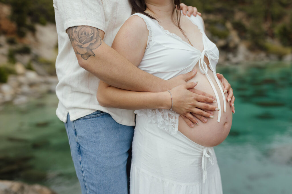 Close up of couple standing together and holding hands on woman's pregnant belly together with Lake Tahoe in background