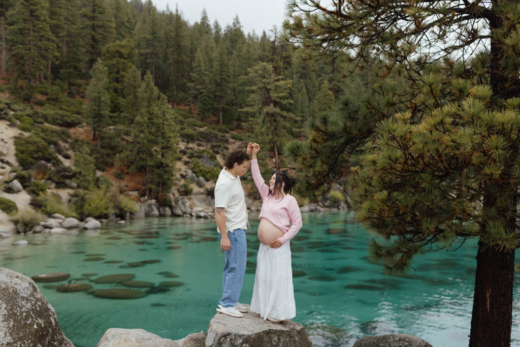 Couple holding hands while standing on rock and woman has hand on pregnant belly with Lake Tahoe in background
