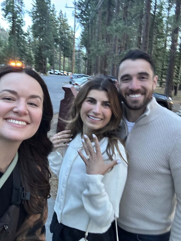 Bridgette Wuest Photographer smiling next to engagement couple while woman shows off engagement ring with pine trees in background at Zephyr Cove