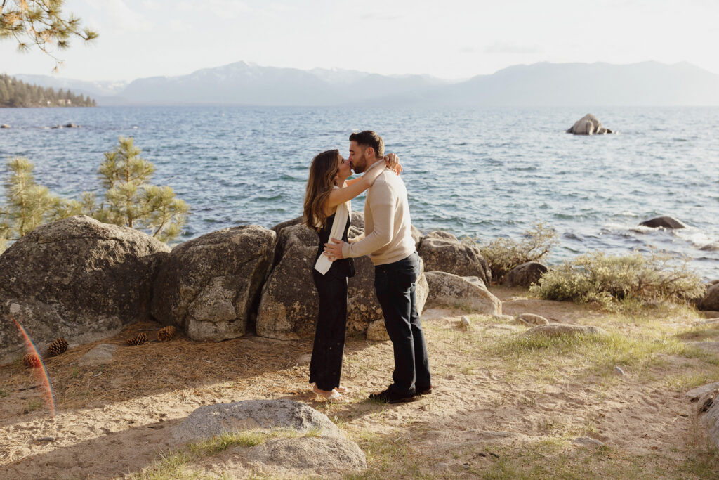 Couple kissing while holding each other and standing in front of large rocks and Lake Tahoe at Zephyr Cove