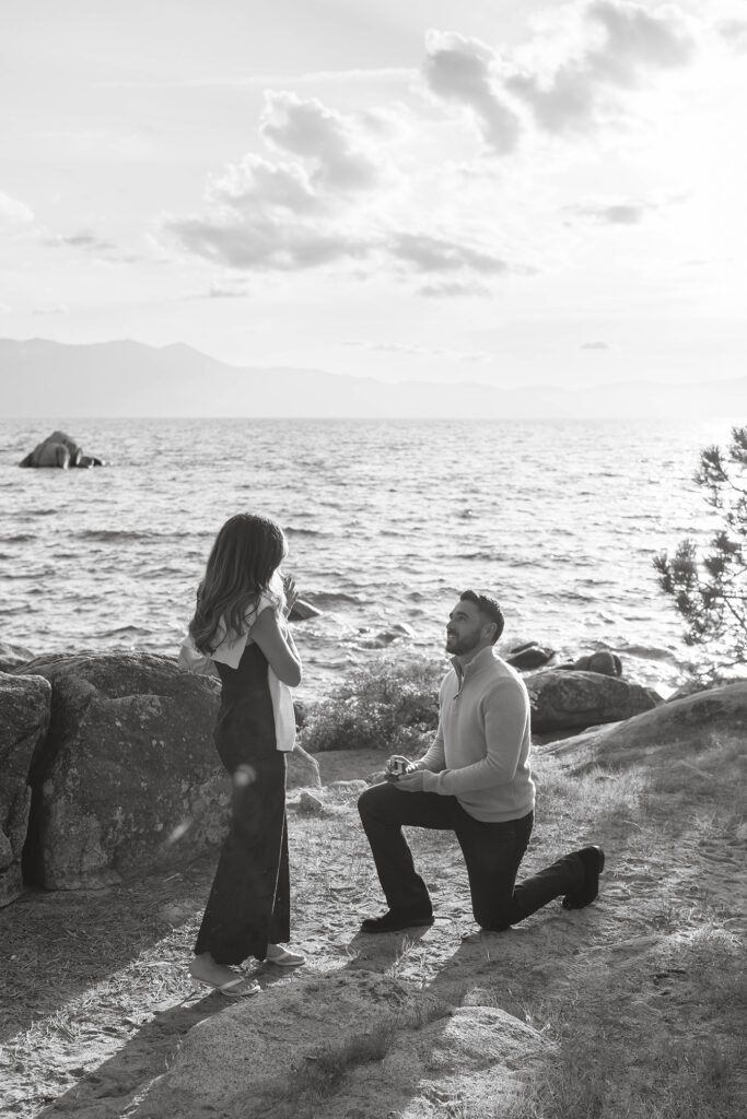 Woman emotional while man on one knee proposes holding engagement ring with Lake Tahoe in background at Zephyr Cove