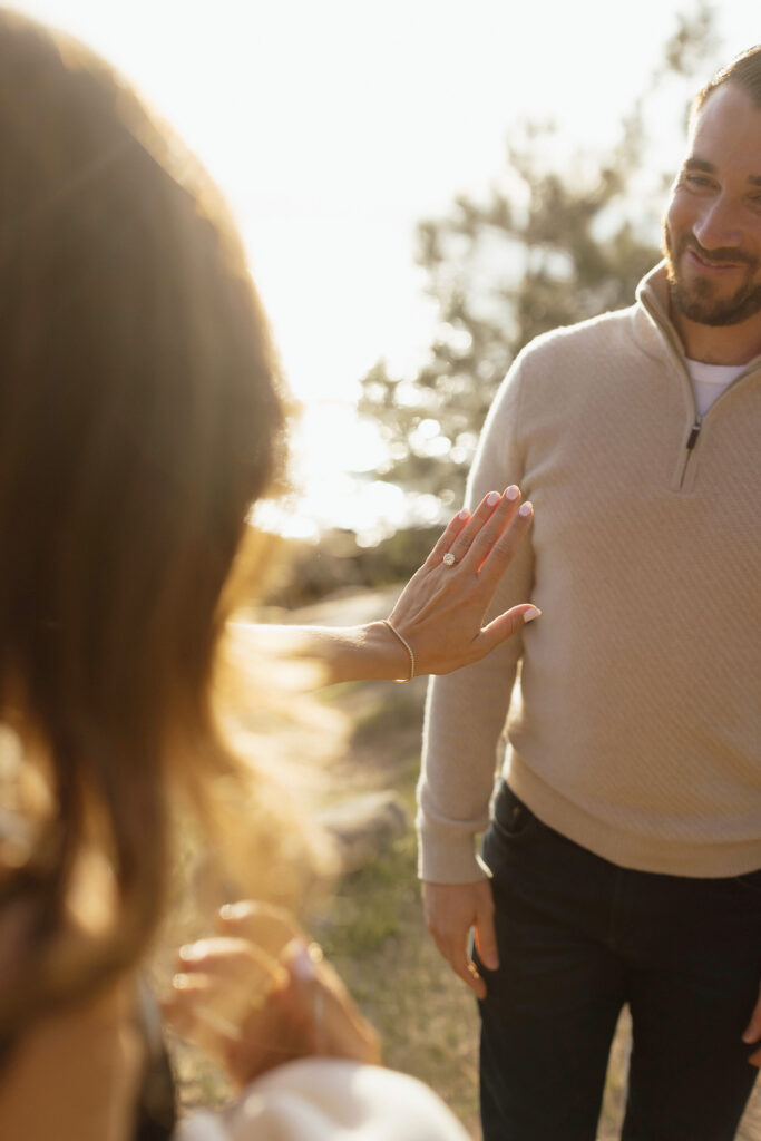 Woman holding out hand and admiring engagement ring while fiancé smiles at her in Zephyr Cove