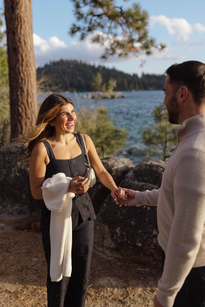 Woman smiling at fiancé while holding hands with him in front of large rocks and Lake Tahoe at Zephyr Cove