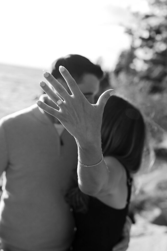 Woman holding hand up close to camera showing off engagement ring while couple kisses behind her hand at Zephyr Cove
