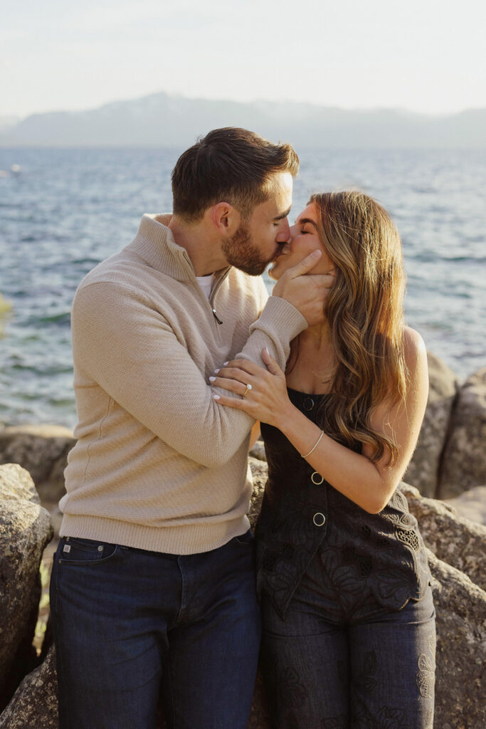 Engagement couple leaning on large rock while holding and kissing each other with Lake Tahoe in background at Zephyr Cove