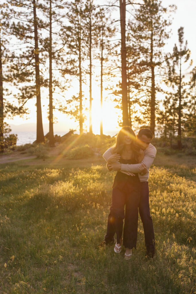 Man hugging fiancé from behind in an open field while she laughs with sunset between pine trees in background at Zephyr Cove