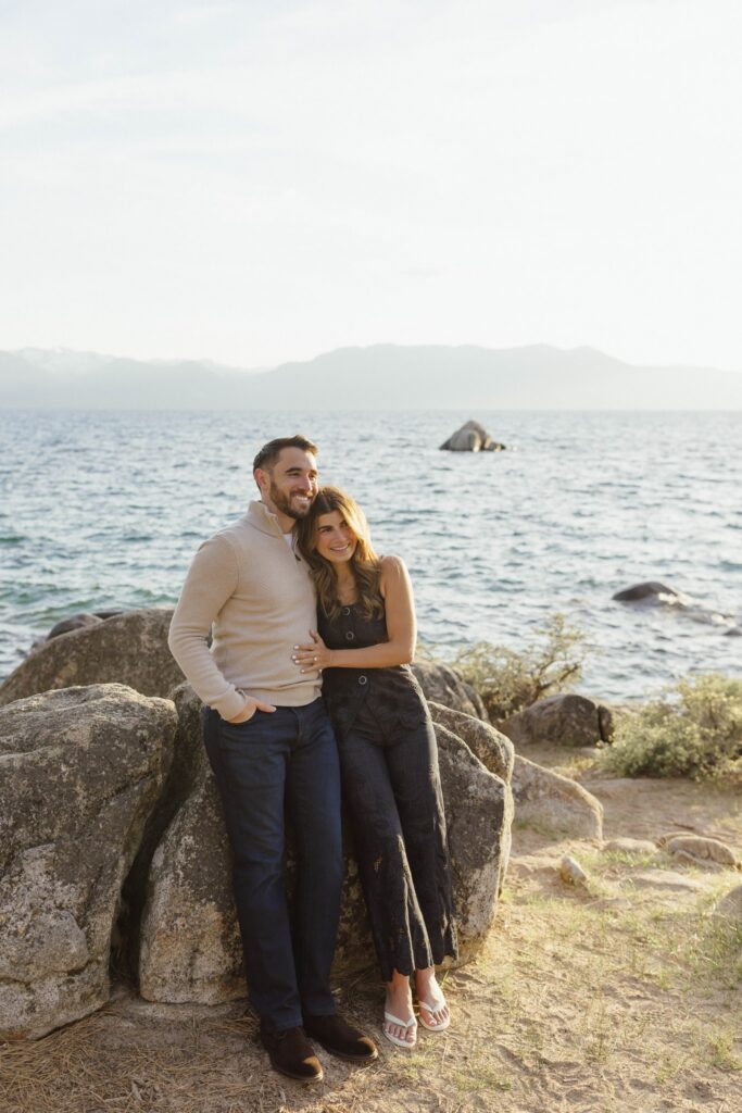 Engagement couple leaning on a large rock together while holding each other and smiling with Lake Tahoe in background at Zephyr Cove